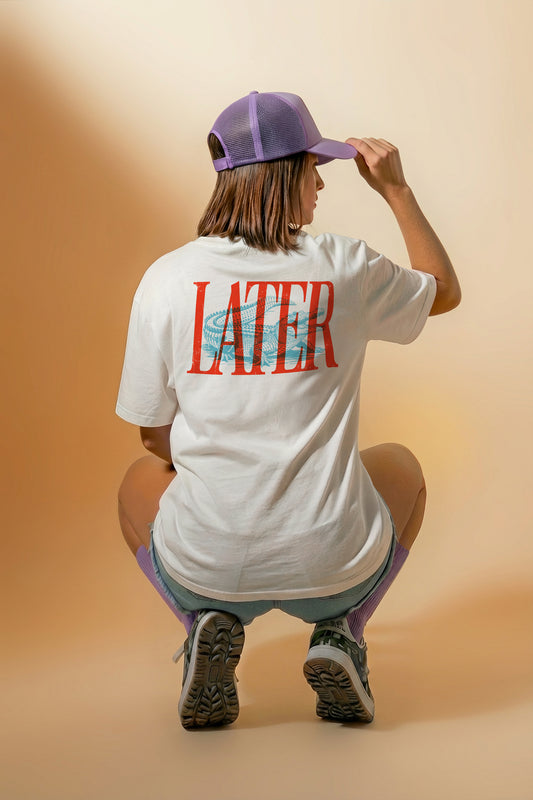 Person wearing a white t-shirt with 'LATER GATOR' printed on the back, sitting on a stool against a beige background.